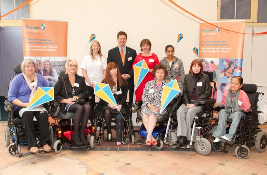 Group of people at a Muscular Dystrophy event, several using wheelchairs, holding colourful signs with charity banners in the background.