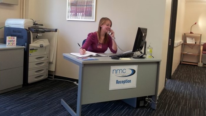 A woman behind a computer desk, talking on the telephone and writing in a notepad.